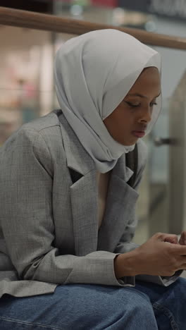 attractive african-american woman with hijab and cellphone rests in shopping mall boutique gallery. black muslim lady in entertainment center. traditions and life