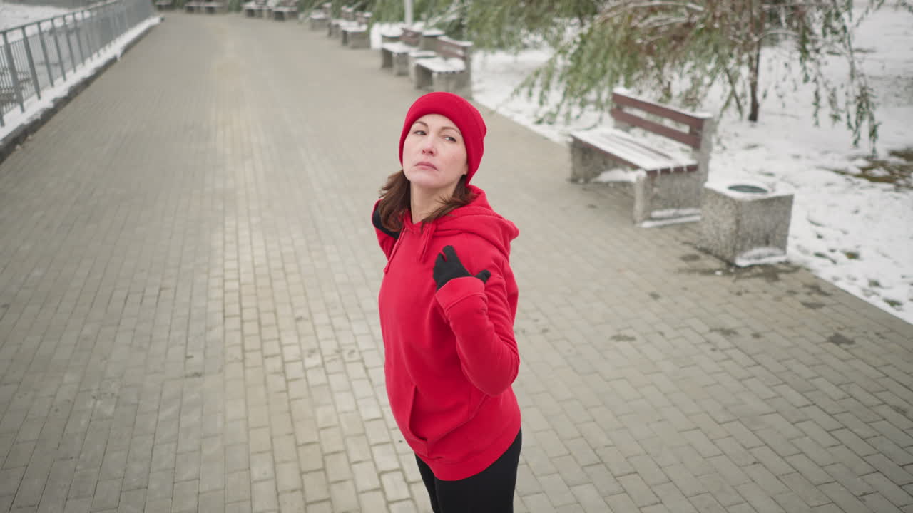 dama realizando estiramiento de manos girando su brazo al aire libre en la rutina de fitness de invierno, vestida con ropa atlética, suelo cubierto de nieve, árboles helados y bancos en un entorno de parque sereno