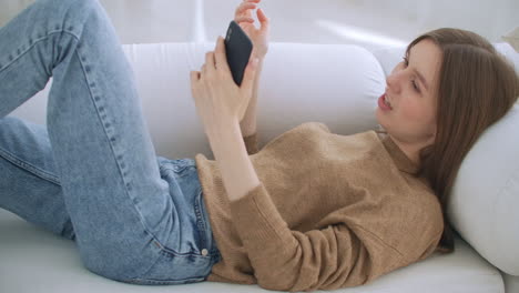 거실의 소파에 누워서 채팅하고 스마트폰을 사용하여 메시지를 쓰고 있는 여성. 외로운 행복한 여성 스마트폰을 사용하여 휴식을 취하고 소파에 앉아서 읽고 쓰고 검색하고 모바일 인터넷에서 검색하고 있습니다.