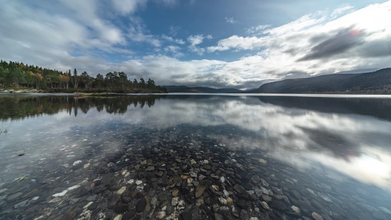 el fondo de un lago poco profundo cubierto de guijarros coloridos se ve a través de las aguas transparentes como nubes rodar por encima y se reflejan en la superficie vidriosa