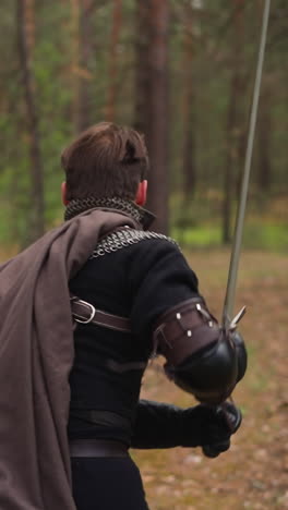 strong knight with cape flying in wind holds sword running through forest backside view slow motion. man takes part in medieval role-playing reconstruction