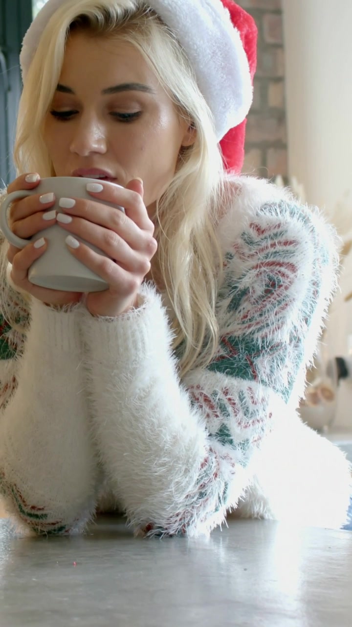 Video verticale: Donna con cappello di Babbo Natale e maglione morbido al bancone, che solleva una tazza e sorseggia per scaldarsi le mani.