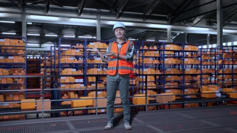 full body of asian male engineer with safety helmet standing in the warehouse with shelves full of delivery goods. smiling and pointing to side recommends about something in the storage