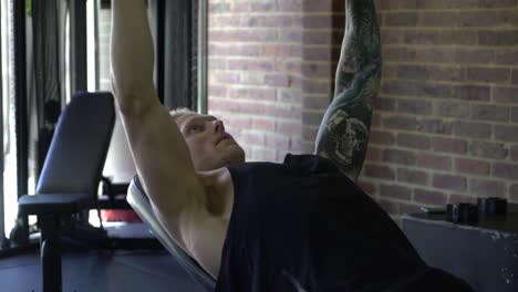 hombre tatuado en el gimnasio en casa de cerca haciendo prensa de pecho inclinada con mancuernas