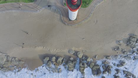 the red and white umhlanga lighthouse on sandy beach with waves crashing on rocks, aerial view