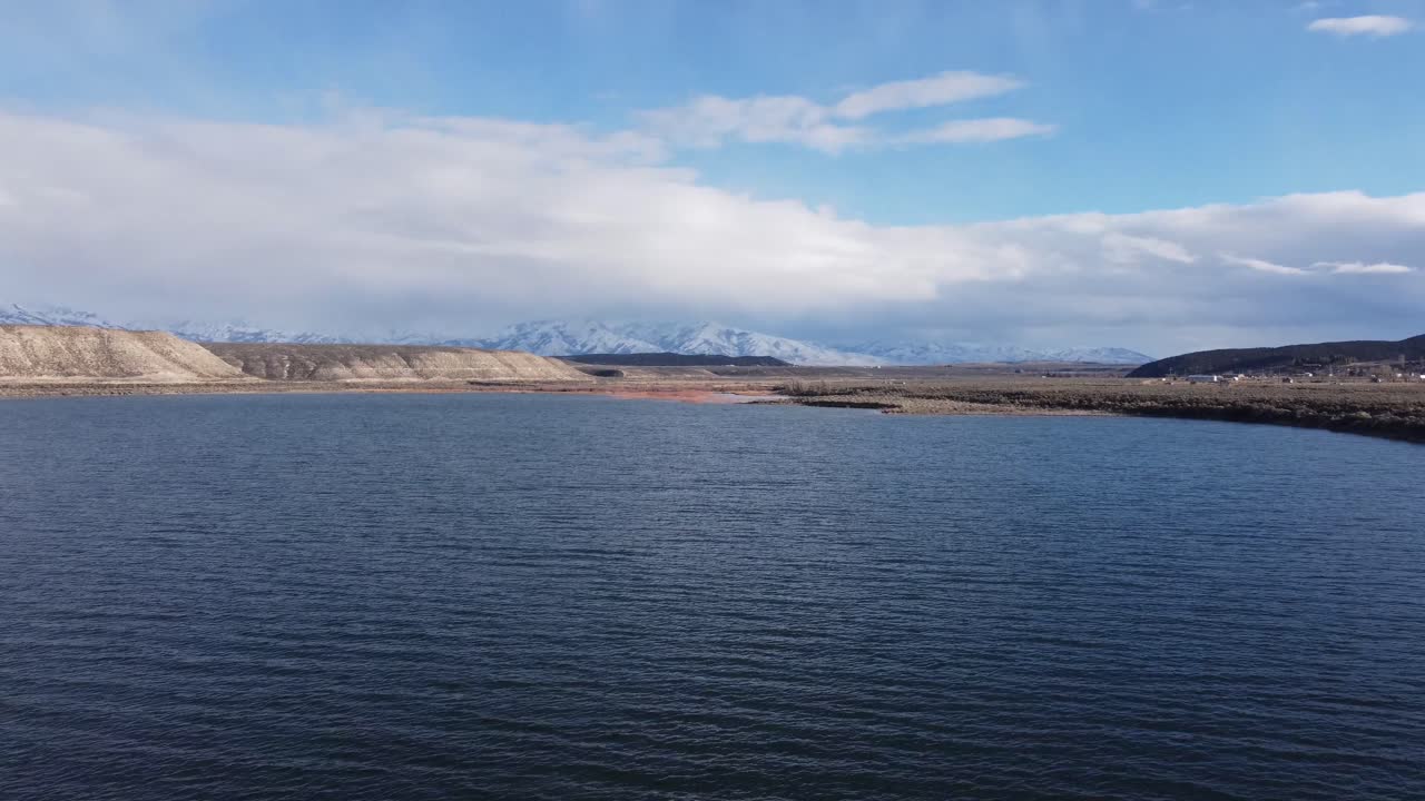 Drone reveal of the snow-covered Ruby Mountain range from the blue water of a lake or river in the Northern Nevada desert. This wilderness winter scene highlights the aerial beauty of the desert.