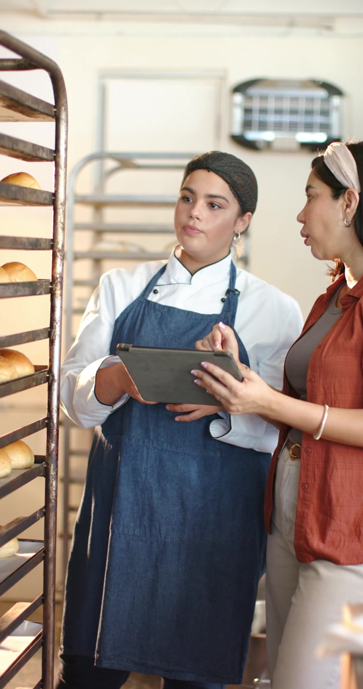 Chef donna e collega diverse con fascia per capelli che controllano pagnotte fresche su una griglia per la qualità, usando un tablet.