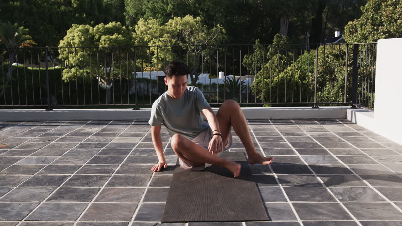 Asian man preparing yoga mat on sunny terrace, enjoying outdoor exercise