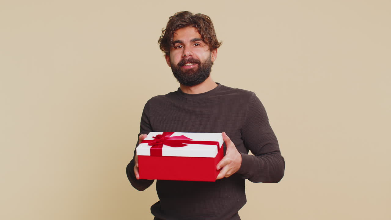 미소 짓는 젊은 인도인 남자가 생일 선물 상자를 제시하고 베이지색 배경에 손을 펴고 있습니다.