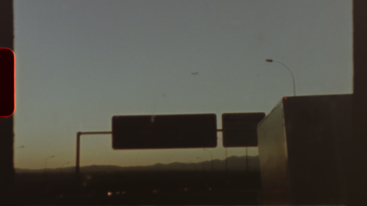 Filmed on super 8 film Grainy vintage 8mm footage, capturing highway stretching across twilight landscape with approaching road signs and distant plane silhouetted against dusky evening sky