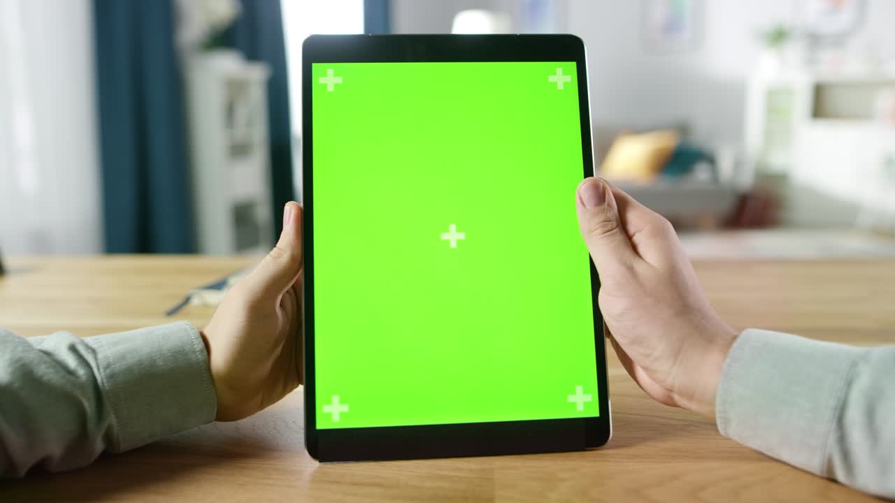 Førstepersonsoptagelse af en mand, der bruger håndbevægelser på en digital tabletcomputer med grøn mock-up-skærm i portrættilstand, mens han sidder ved sit skrivebord. I baggrunden hyggelig stue