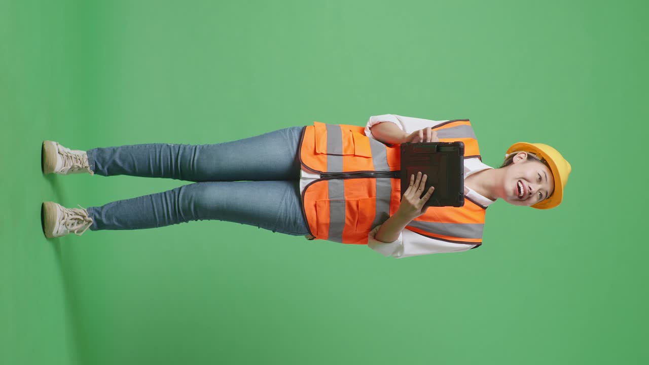 Full Body Of Asian Female Engineer With Safety Helmet Looking At The Tablet In Her Hand And Looking Around While Standing In The Green Screen Background Studio