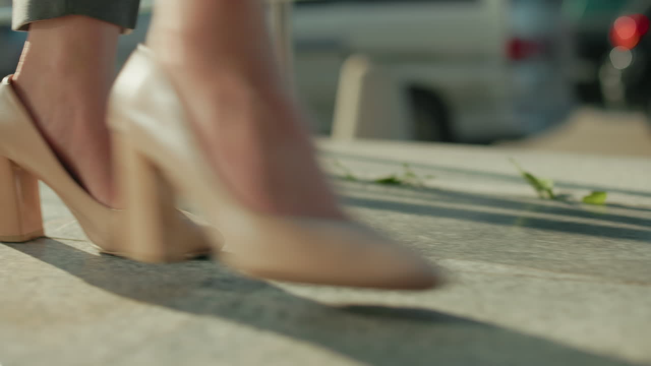 Side view of lady in heels walking by iron railing casting long shadow on ground with blurred parked and moving cars in background under bright sunlight