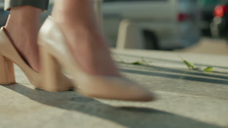 side view of lady in heels walking by iron railing casting long shadow on ground with blurred parked and moving cars in background under bright sunlight