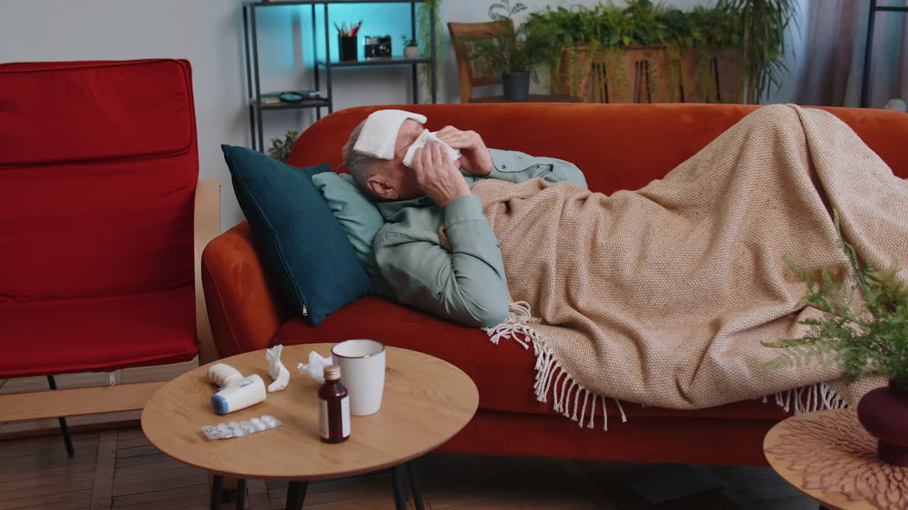 Sick ill elderly man suffering from cold allergy lying on home sofa sneezes wipes snot into napkin