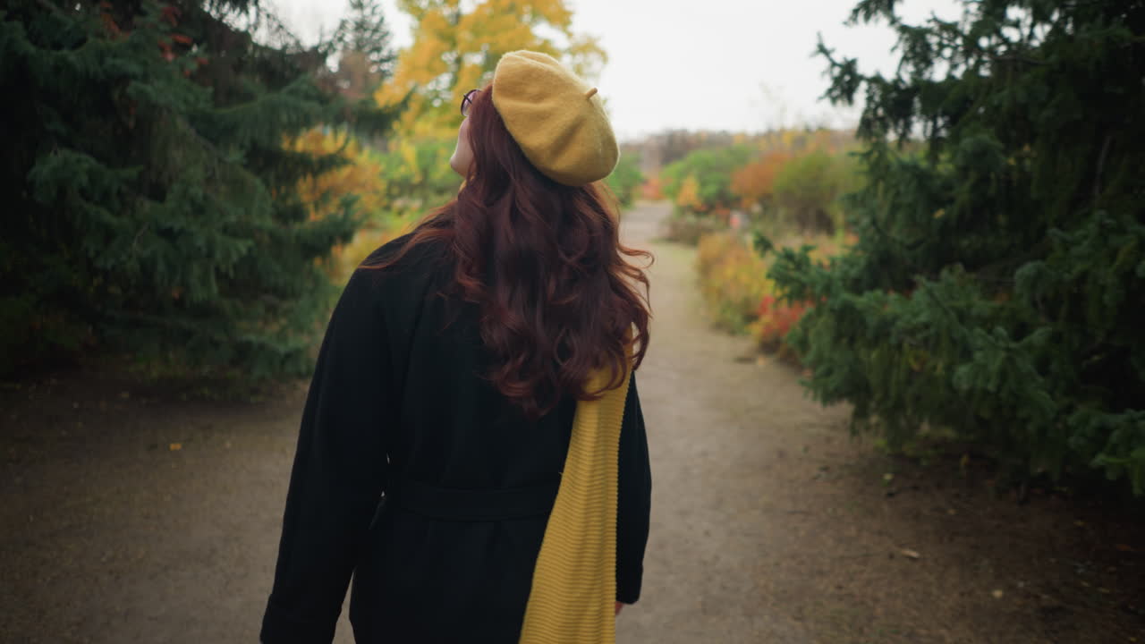Bakfra-bilde av en dame i gul beret og matchende skjerf, som tar en høstspasertur i en hage, ser seg rundt og justerer sitt røde bølgete hår mens hun går.