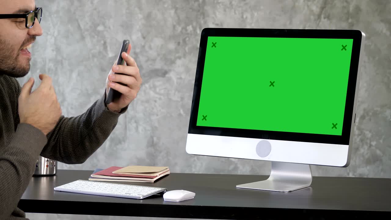 atractivo hombre de barba revisa sus dientes en sus teléfonos cámara selfie cerca del monitor de la computadora. pantalla verde muestra de maqueta