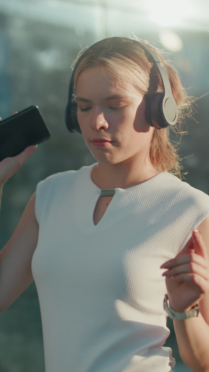 White lady in white top holding smartphone and wearing headphones dances joyfully outdoors with sunlight illuminating modern residential building and parked cars in urban setting