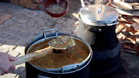 a female sewer pours soup into a clay mold bowl with a wooden spartle from a large saucepan outdoor