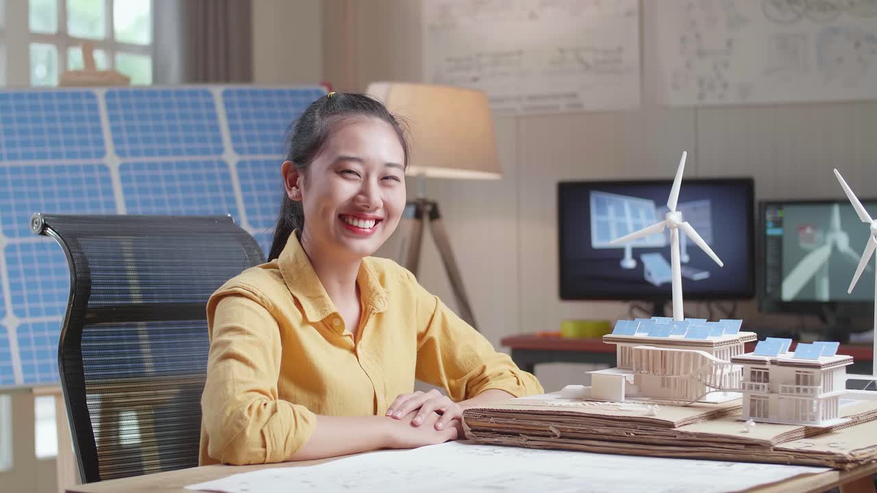 태양 전지 패널 을 가진 작은 집 의 블루프린트 와 모델 을 작업 하는 동안 아시아 여자 가 카메라 에 미소 짓는다