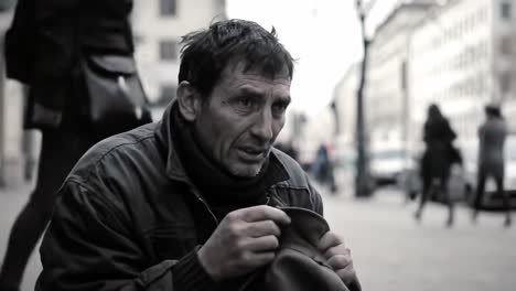 a man sits on a busy city street looking introspective and seeking assistance. he clutches a simple hat, surrounded by passing pedestrians.