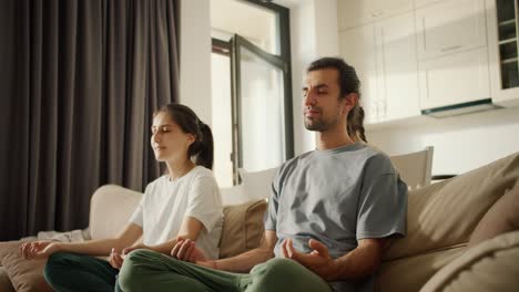 A-brunette-girl-in-a-white-T-shirt-together-with-her-husband,-a-brunette-man-in-a-gray-T-shirt,-meditates-while-sitting-on-light-brown-daughter-girl,-a-little-brunette-girl-in-a-yellow-dress-interferes-with-them-and-has-fun-in-a-modern-studio-apartment