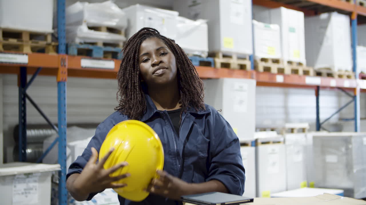 trabajadora afroamericana quitándose el casco, apagando la tableta y sonriendo a la cámara en el almacén