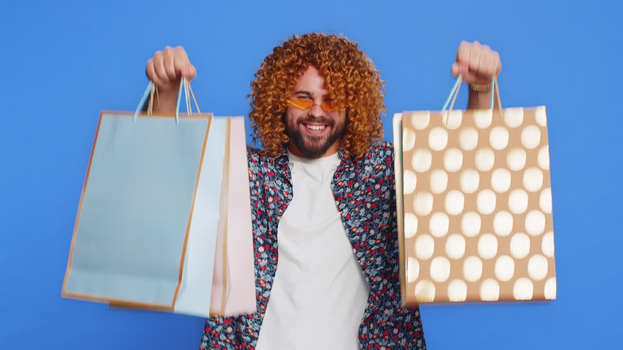 hombre caucásico mostrando bolsas de compras anunciando descuentos sonriendo pareciendo asombrado por los bajos precios
