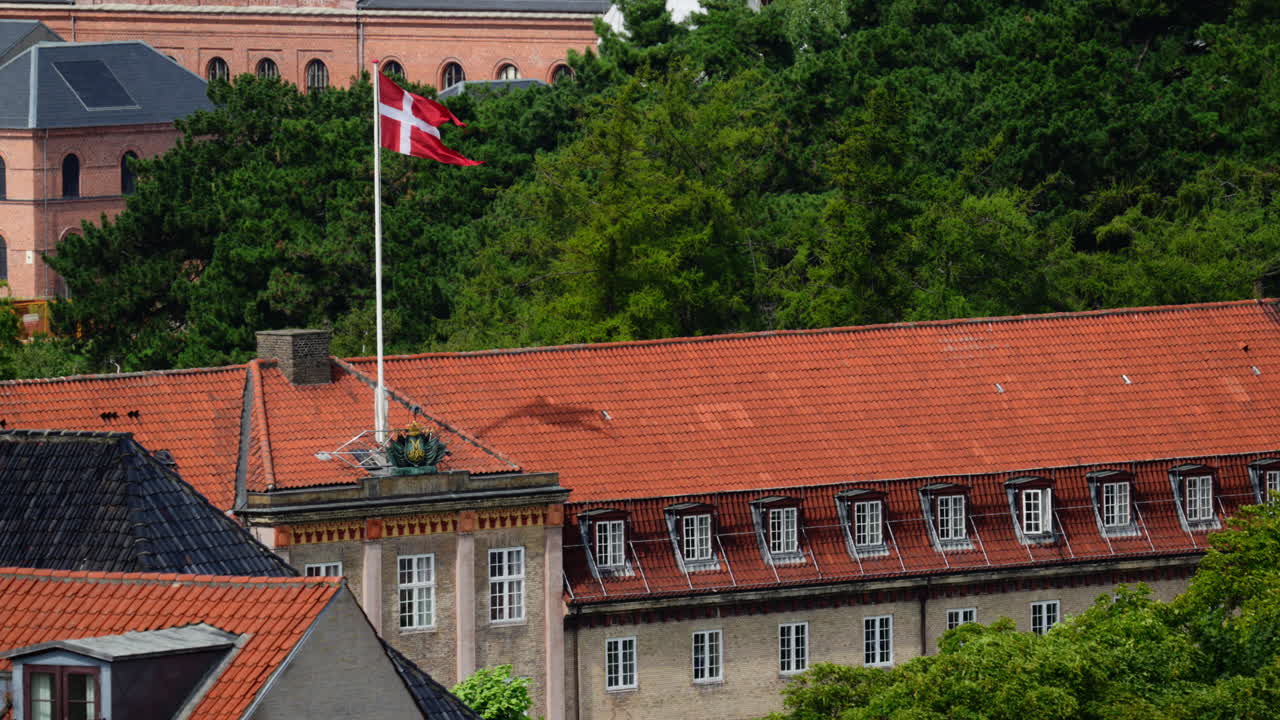 Dansk flag, der vajer på en flagstang over en bygning med et rødt tegltag og en grøn baggrund med træer