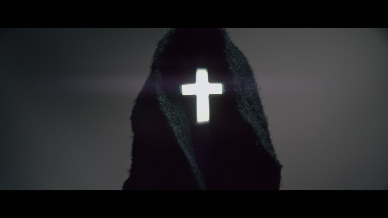 Masked and hooded man with cross of light glowing on his face looks in the camera