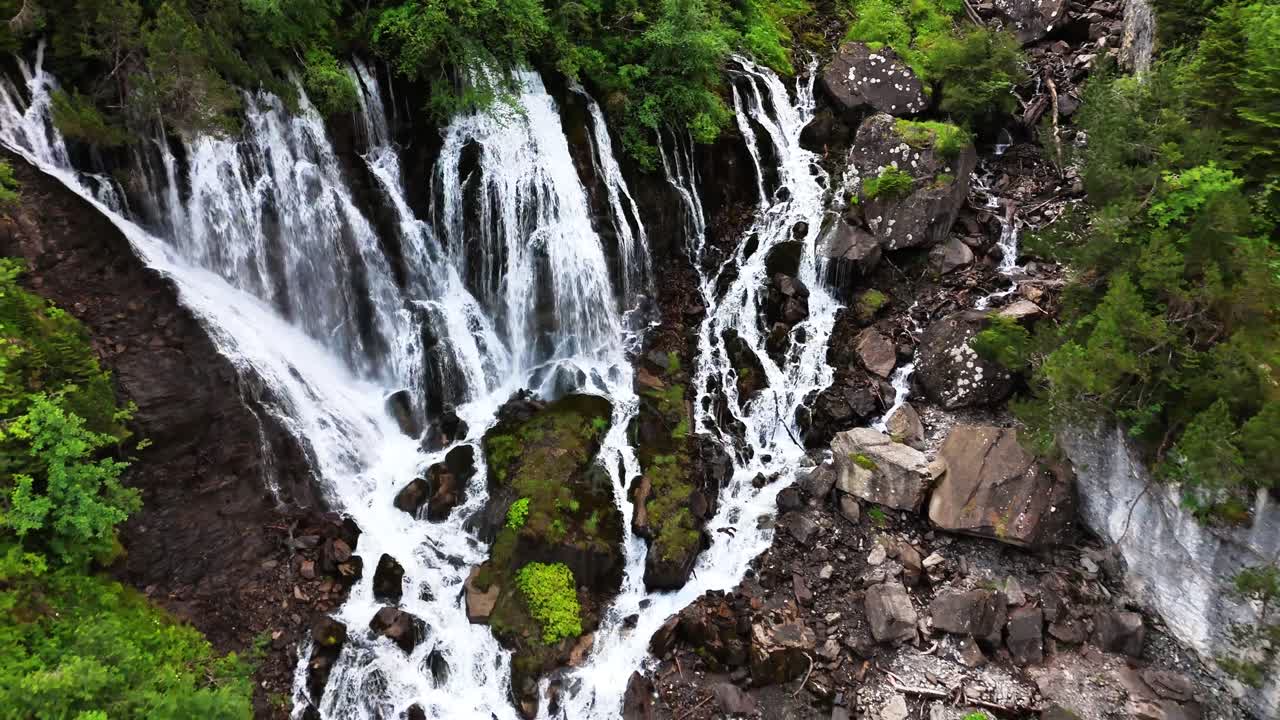Cascading waterfall at Sibenfälle in Lenk, Switzerland, flowing over mossy rocks and rugged terrain surrounded by lush green forest and steep cliffs, creating a striking alpine natural landscape