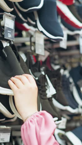 dama de vestido rosa en la tienda de zapatos examina una zapatilla negra, observando su suela de cerca, el estante delante de ella muestra más zapatos en varios estilos, ligeramente borroso, con la tienda bien iluminada