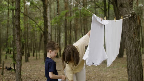 caucasian woman hanging out washing clothes on clothesline with the help of her son at the camping in the forest