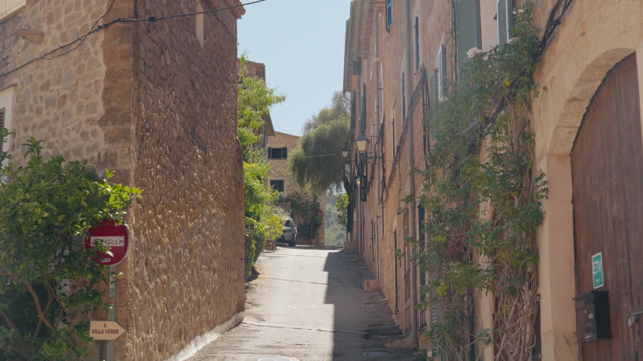 Ampia inquadratura di una stretta via illuminata dal sole tra case in pietra dorata ricoperte di piante rampicanti. Atmosfera di un tranquillo villaggio mediterraneo a mezzogiorno