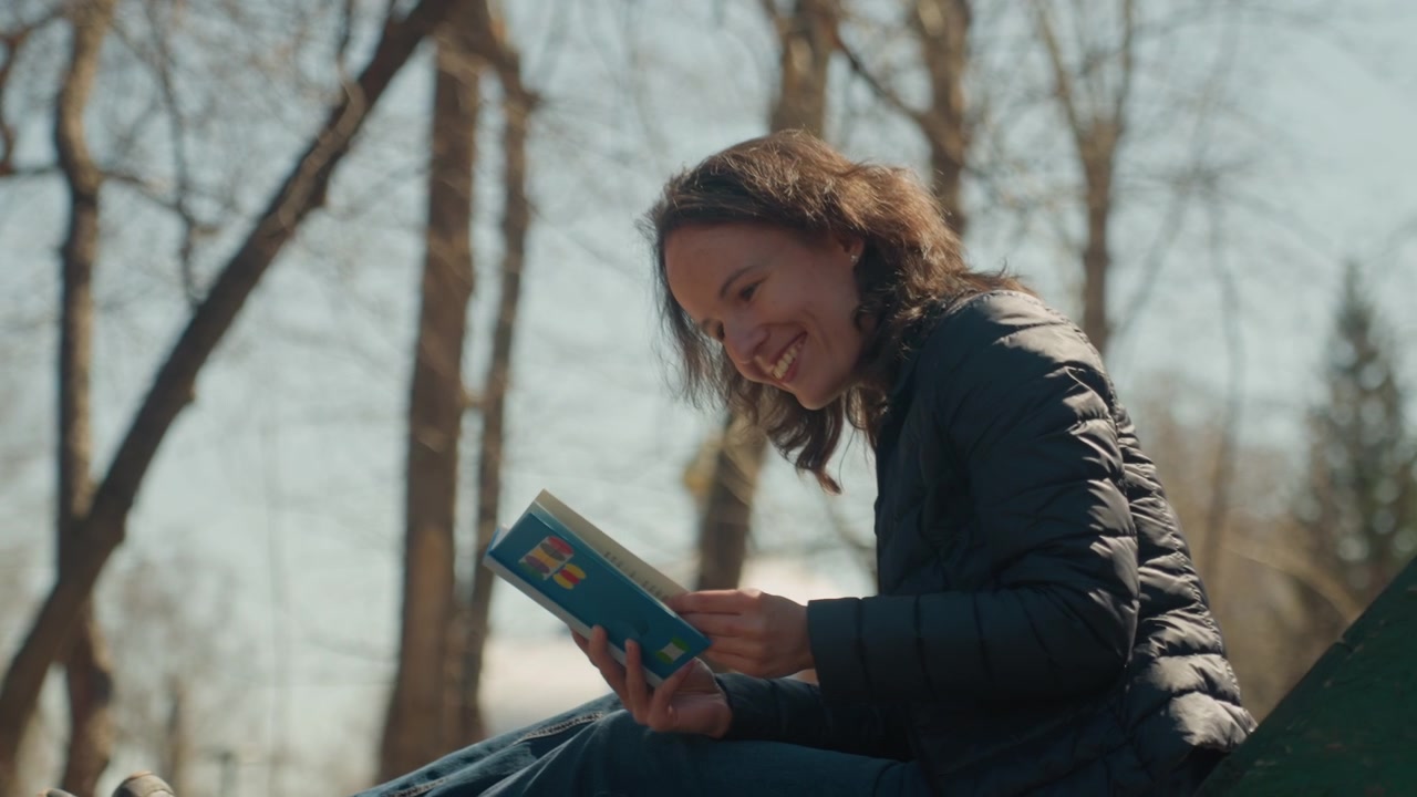 Close up of woman reading open book while seated outdoors on green bench. Woman wears dark puffer jacket and blue jeans with bare trees visible in soft daylight background. Book is held