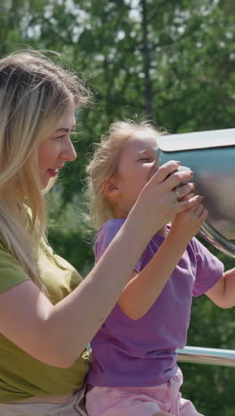 careful mother helps curious little girl look through large binoculars on viewing ground at highland resort on sunny day slow motion