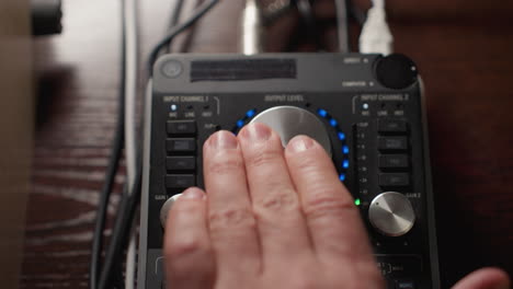 close-up of a hand operating a sound mixer, rotating a large knob to the left to adjust audio levels. with the mixer s illuminated interface and various controls visible