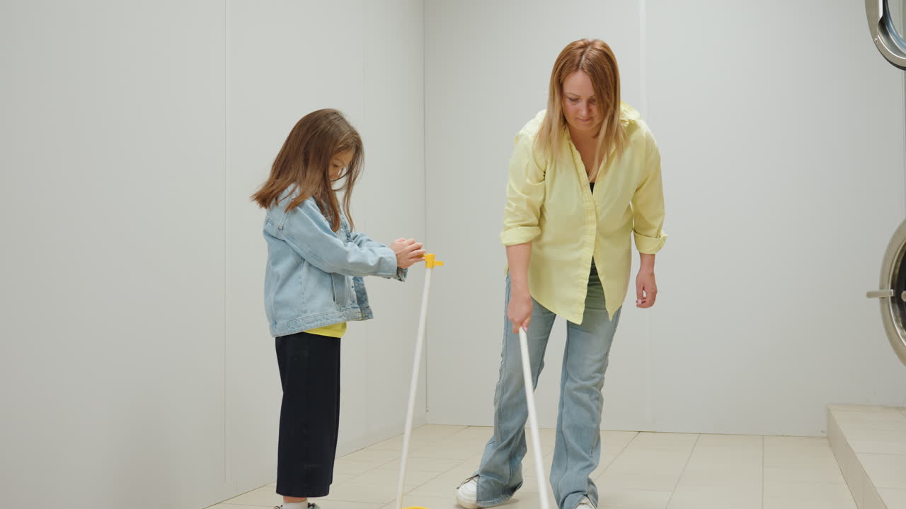 Zoom in on nurturing mother sweeping tiled floor while little girl holds dustpan, cooperative cleaning inside laundromat by industrial washers, family teamwork, guidance, hygiene, stainless interior