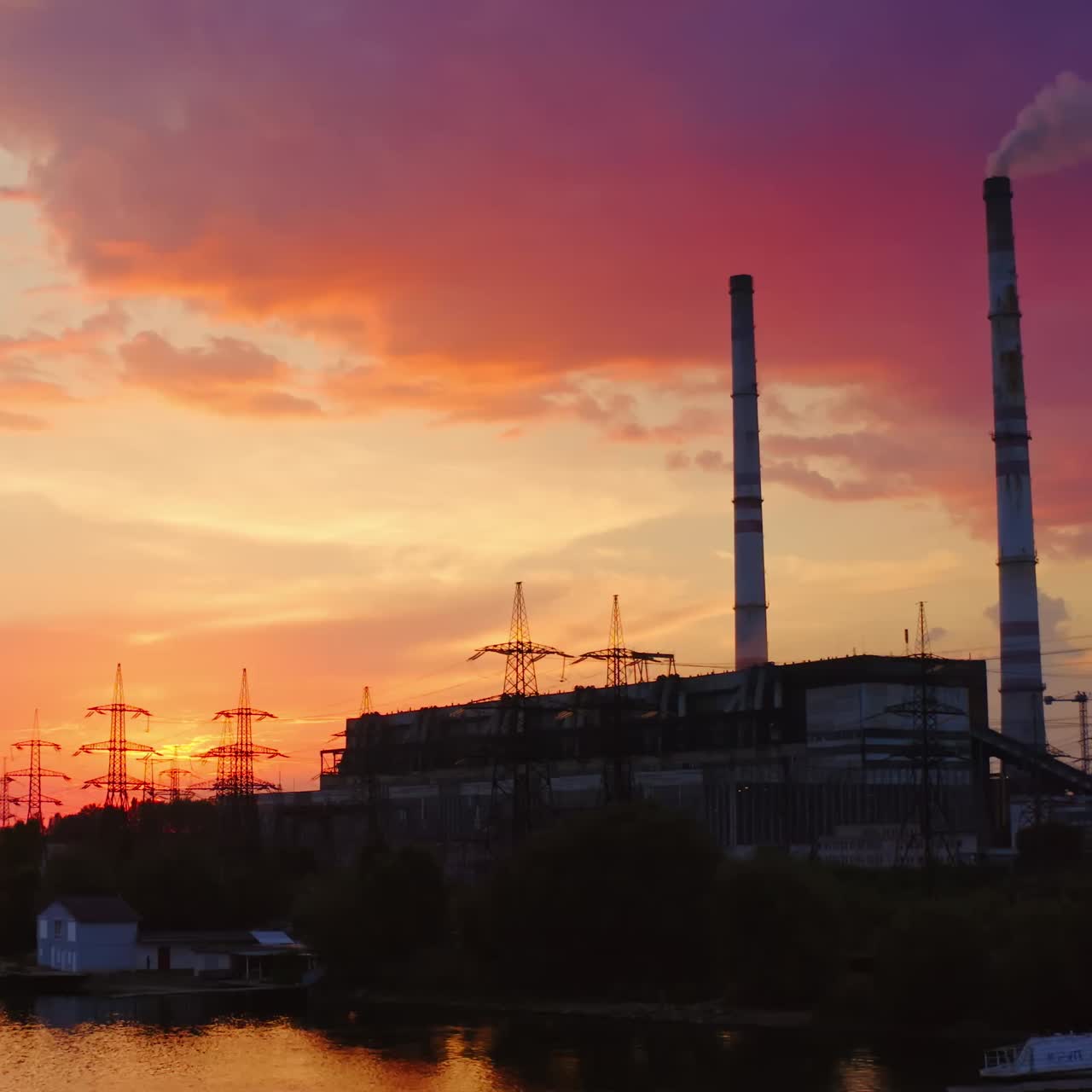 Industrial zone in the evening nature. Transmission lines, factory with pipe releases smoke near the river at sunset. View from the river