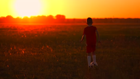 Kleiner-Fußballspieler-In-Einem-Roten-T-Shirt,-Der-Bei-Sonnenuntergang-Mit-Dem-Ball-über-Das-Feld-Läuft,-Zur-Sonnenkamera-Auf-Steadicam