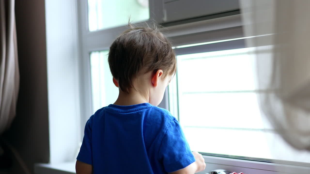 Dark-haired baby boy stands at the window. Toddler sticks a toy to the glass and spins it turning to camera.