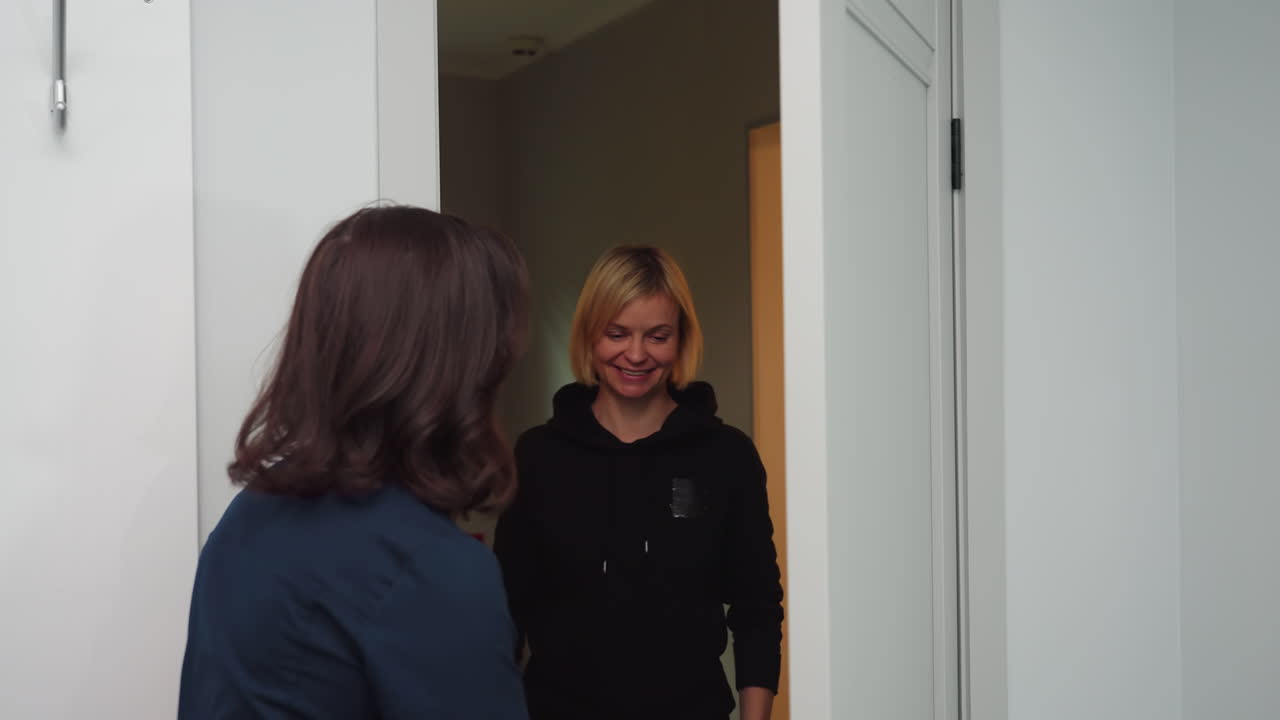 Massage therapist in navy uniform opens door with warm smile to welcome client who smiles back in bright white treatment room, creating friendly, professional atmosphere before relaxation session