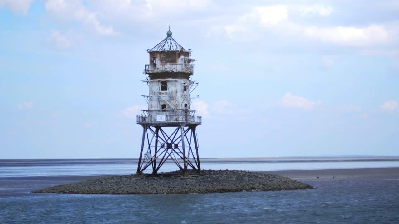 Cormorans flying around and nesting on an old abandoned lighthouse called cormoran tower standing in the wadden sea with water flowing beneath and a cloudy sky. Camera passing by from a boat.