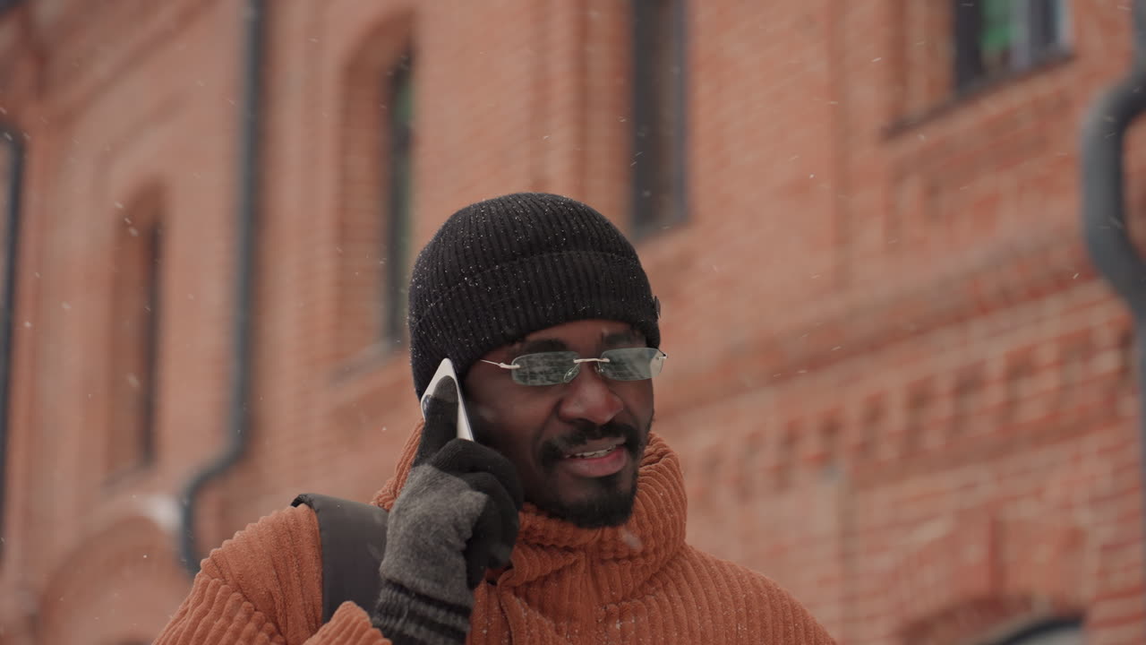 Svart mann smiler i telefonen, går forbi en mursteinsfasade med snøflak på lue og briller, vennlig urban stemning. Første vinkel viser en entreprenør som avslutter en avtale over telefon, andre vinkel fanger en influencer.
