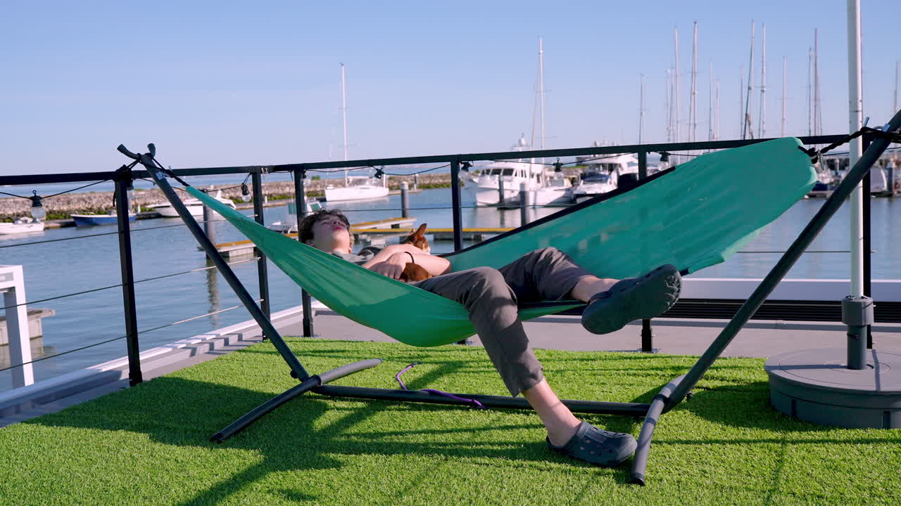 Teenager relaxing in teal hammock with small dog, overlooking marina serene waterfront on bright sunny day, embodying peaceful outdoor lifestyle and leisure moment