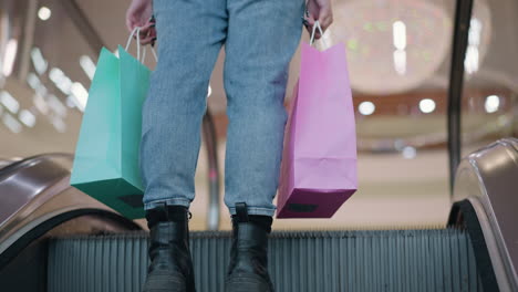 leg view of woman with two shopping bags on moving escalator in mall, steps off onto brown rug with blurred background of mall's interior