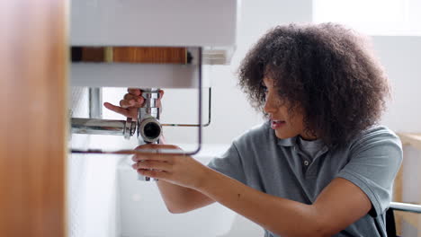 young black female plumber sitting on the floor unscrewing the trap pipe under a bathroom sink, seen from doorway