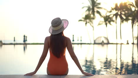 Vista-Trasera-De-La-Mujer-En-Traje-De-Baño-Naranja-Sentada-Al-Borde-De-La-Piscina-En-Un-Hotel-Exótico-En-Hawaii-Al-Atardecer