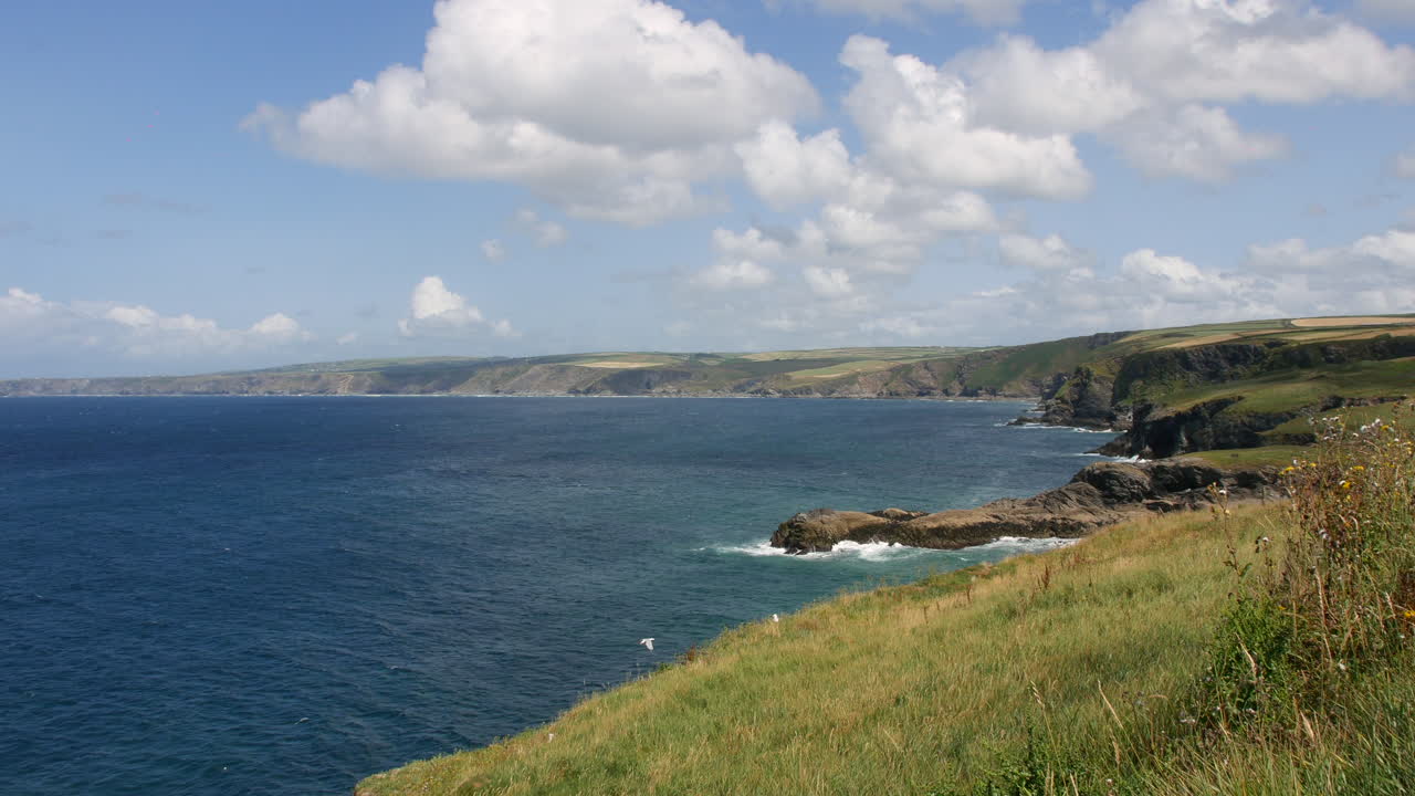 Cliffs overlooking the ocean are stretching along the rugged coastline of Cornwall near Port Isaac and Portwenn, with waves crashing below and rolling hills in the distance