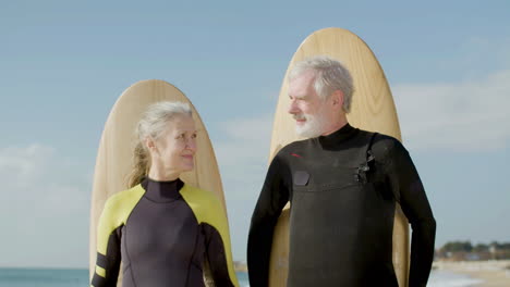 front view of a senior couple with surfboard behind their backs standing on the ocean shore and looking ath the camera